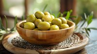 Fresh green olives in wooden bowl on woven placemat with olive leaves, natural light setting creates appetizing Mediterranean food composition.