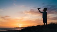 Young boy silhouette holding toy airplane against dramatic sunset sky over water. Child playing outdoors during golden hour, suitable for childhood, imagination, dreams, and adventure themes.