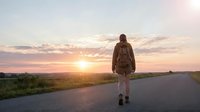 Young woman with brown hair wearing a backpack and athletic clothing standing on a rural road during golden hour sunset, facing away from camera, adventure and travel concept.