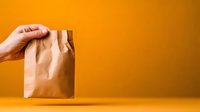 Caucasian hand holding brown kraft paper bag against vibrant orange background, minimalist composition for delivery and takeout concepts.