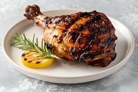 Grilled chicken leg quarter with honey glaze and charred marks served on white ceramic plate with fresh rosemary sprig and grilled lemon slice on light background.