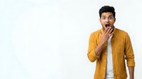 Young man with dark hair and South Asian ethnicity wearing a mustard yellow button-up shirt over a white undershirt, displaying a shocked or surprised facial expression with hand near mouth against white background.