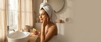Young woman with towel on head applying skincare product in modern bathroom with natural sunlight for morning beauty routine and self-care wellness.