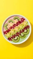 Smoothie bowl topped with sliced kiwi, star fruit, and pomegranate seeds on bright yellow background. Healthy breakfast or dessert option.