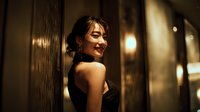 Young Asian woman in elegant black dress smiling confidently in upscale restaurant or hotel lobby with warm ambient lighting and modern interior design.