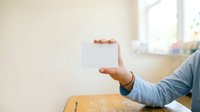 Man in blue shirt holding blank white card at wooden table, suitable for business messaging, advertising mockups, and corporate communications.
