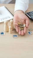 Hand holding coins with wooden family figures and insurance icons representing financial protection planning and budget management for household expenses.