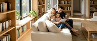 Happy family of three reading together on beige sofa in modern home library with wooden bookshelves, perfect for lifestyle and education content.