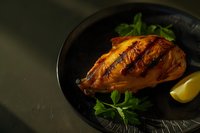 Grilled chicken breast with golden-brown grill marks served on black plate with fresh parsley and lemon wedge against dark background. Moody food photography.
