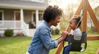 African American mother pushing laughing toddler daughter on backyard swing, sharing joyful moment outdoors near family home on sunny day.