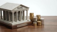 Classical bank building model with golden coins stacks on wooden surface representing financial services, investment growth and monetary system concepts.