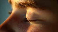 Close-up macro shot of young Caucasian person's eye area with freckles and long eyelashes in warm golden sunlight showing skin texture and natural beauty.