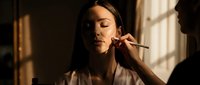 Young woman applying makeup with brush in warm natural light by window creating intimate beauty routine moment for cosmetics and self-care content.