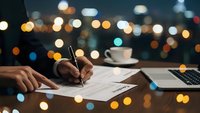Professional businessman writing documents at desk with pen during evening hours with city lights bokeh background creating productive workspace atmosphere.