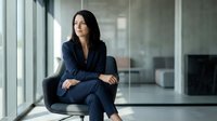 Confident businesswoman in navy suit sitting in modern office space with glass walls and natural lighting for corporate and professional concepts.