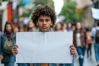Young African American male activist with curly hair holding blank white protest sign during street demonstration, crowd in background, serious expression.