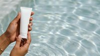 Close-up of hand holding blank white sunscreen tube against rippling water background. Skincare product mockup for sun protection advertising and cosmetic branding campaigns.