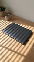 Closed laptop with black padded case on wooden desk beside ceramic mug and potted plant, bathed in natural sunlight casting striped shadows for workspace and office design projects.