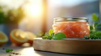 Fresh salmon pieces in glass jar with basil leaves and citrus on wooden board, soft natural lighting creates appetizing mood for food photography.