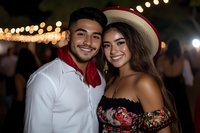 Young Hispanic couple enjoying outdoor festival with string lights, man in white shirt with red bandana, woman in floral top and cowboy hat smiling together.