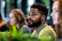 Young African American man with beard and curly hair wearing yellow sweater listening attentively during business meeting or conference presentation.