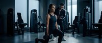 Young athletic woman performing lunges with dumbbells in modern gym while male trainer observes her form during strength training workout session.