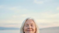 Senior woman with white hair smiling with eyes closed against serene ocean and sky backdrop at sunset, conveying peace and contentment.