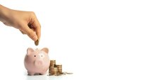 Hand dropping coin into pink piggy bank with stack of golden coins on white background for savings and financial planning concepts.