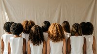 Diverse group of women showing various natural hair textures and styles from behind, wearing white tops against neutral backdrop for beauty concepts.
