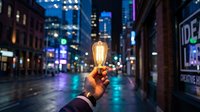 Hand holding glowing vintage Edison bulb on blurred city street at night with colorful neon lights and urban bokeh background for creative business concepts.