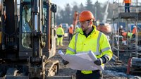 Construction worker in safety gear reviewing blueprints at active job site with heavy machinery and crew in background for infrastructure projects.