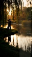Romantic couple silhouette embracing by tranquil lake at golden hour sunset with weeping willow tree branches and warm light reflections on water.