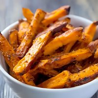 Crispy baked sweet potato fries seasoned with herbs and spices served in white ceramic bowl on rustic wooden table, selective focus on golden-brown edges.
