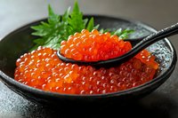 Fresh red caviar served in black ceramic bowl with spoon and green garnish. Bright orange salmon roe closeup showing delicate texture and glossy spheres.