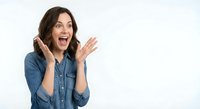 Excited young Caucasian woman with brown hair in denim shirt talking on smartphone with surprised expression and raised hand isolated on white background.