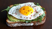 Sunny side up egg on avocado toast garnished with fresh herbs, red pepper flakes and black pepper, served on rustic bread against dark background.