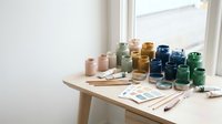 Artist workspace with colorful paint jars and brushes on wooden table by window for creative projects and art supplies organization.