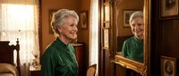 Senior Caucasian woman with gray hair in elegant green dress looking at ornate mirror reflection in vintage Victorian interior with warm lighting.