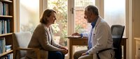 Middle-aged Caucasian woman consulting with African American male doctor in bright medical office with bookshelves for healthcare communication.