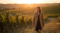 Young woman in beige coat standing in vineyard at golden hour sunset with rolling hills landscape background for lifestyle and travel content.