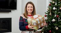 Smiling middle-aged Caucasian woman in festive Christmas sweater holding wrapped gift boxes near decorated tree in cozy living room during holidays.