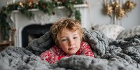 Young child with curly hair wrapped in cozy gray knit blanket wearing festive red pajamas, relaxing by decorated Christmas fireplace.