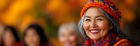 Smiling senior Asian woman wearing colorful traditional headscarf and clothing at outdoor cultural festival with warm autumn background.