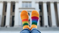 Colorful rainbow striped socks against blurred classical building background, feet up in air showing vibrant orange, pink, yellow, green and blue stripes in playful pose.
