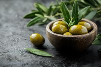 Fresh green olives served in rustic wooden bowl with olive leaves on dark stone background, Mediterranean cuisine ingredients for gourmet cooking.