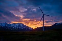 Wind turbine silhouetted against dramatic sunset sky with snow-capped mountain range, highlighting renewable energy in pristine wilderness landscape at dusk.