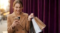 Happy young Caucasian woman in beige coat checking smartphone while holding shopping bags outdoors, smiling at mobile phone screen during retail therapy.