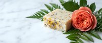 Handmade soap bar with dried flowers and herbs next to coral rose and green fern leaves on marble surface for spa, wellness, and natural skincare product marketing.
