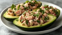 Stuffed avocado halves filled with tuna salad mixed with fresh herbs and vegetables, served on white plate for healthy meal preparation.