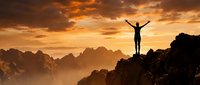 Person celebrating success on mountain peak at golden sunset with arms raised in triumph over dramatic landscape.